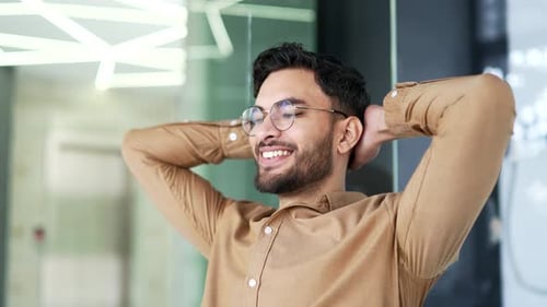 Happy businessman relaxes with hands behind head sitting at workplace in business office.