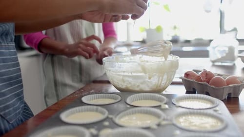 Midsection of biracial mother and daughter baking, preparing cake mix in kitchen, slow motion