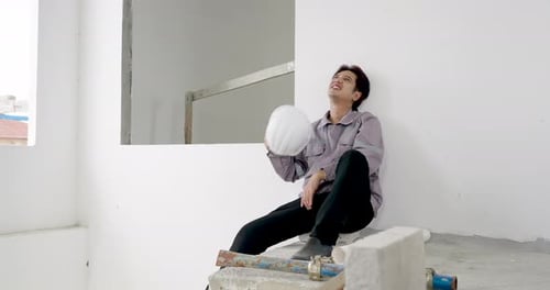 Asian male worker sits in slow motion holding hard hat during construction site safety inspection,