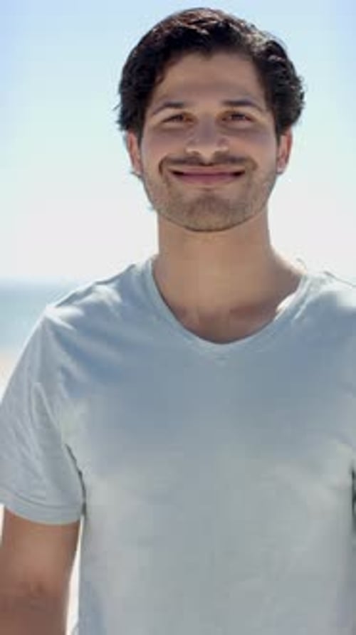 Vertical video: Smiling man enjoying sunny day at beach, wearing casual t-shirt