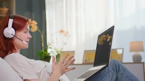 Call Center Beautiful Woman with Headset Talking on Video Call on Laptop While Lying on Bed in Room