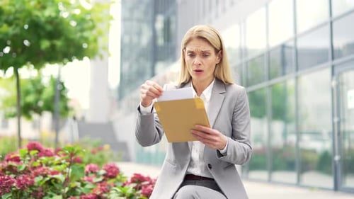 Disappointed businesswoman reading letter with bad news sitting on a bench on street near an office