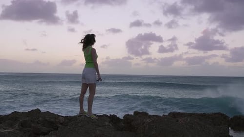 Silhouette Woman at Dusk Raising Arms in Air at Ocean Waves Crashing on Rocks