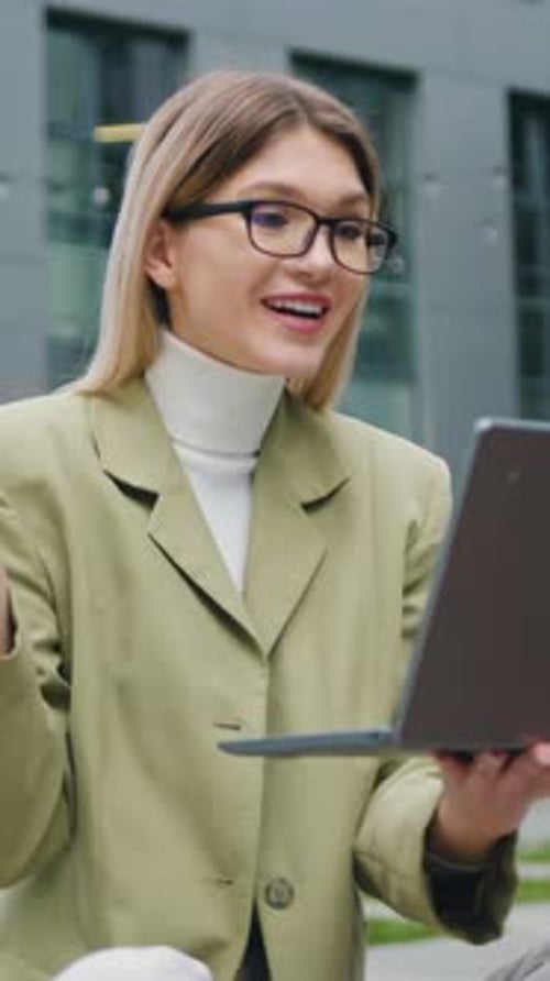 Outdoors Portrait of Smiling Girl in Glasses Having Conference Call on Laptop with Sitting on Street