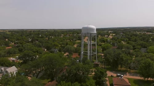Wide aerial shot of a small woodland town centered on a white water tower as the drone performs a