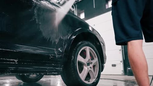 A male car wash employee applies car wash detergent to a black luxury car using spray gun in the car