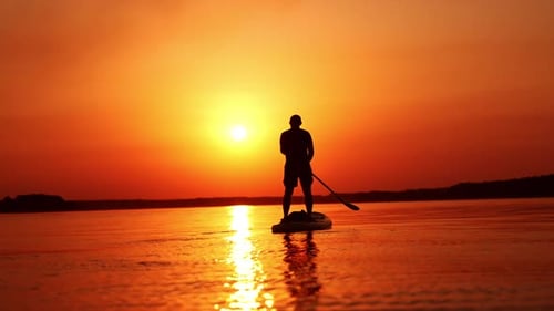 Man stands on the sup board facing the sunset. Man holds a paddle and row with it from each side.