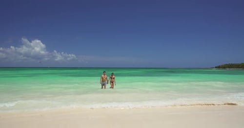 Attractive caucasian couple walking out of turquoise ocean waves on tropical beach
