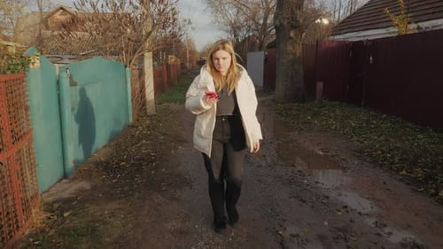 A Young Woman Strolls Along a Muddy Road with Old Deteriorating Houses Around Her Using a Smartphone