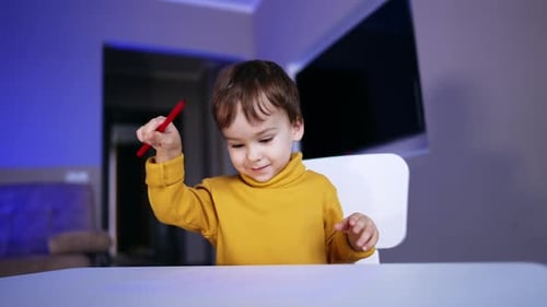 Schönes Kleinkind in gelbem Pullover sitzt am Schreibtisch und zeichnet.