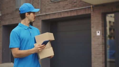 Male Courier Going Along Building Holding the Paper Boxes and Mobile Phone in Hand Delivery Man