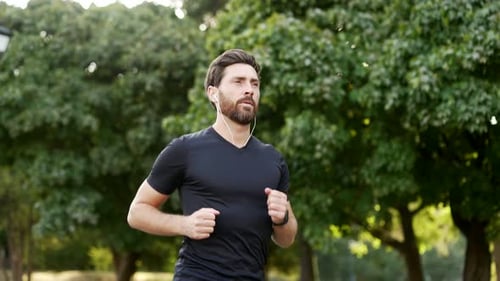Handsome male runner jogging in an urban city park and looking smart watch. Bearded athlete using