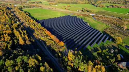 Aerial view approaching a sunlit solar energy farm, in middle of fall foliage