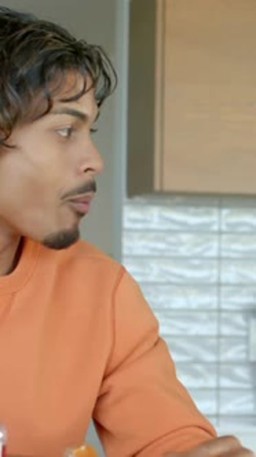 Vertical video: Eating man in orange top hearing knock, turning right at kitchen, sandwich bottle