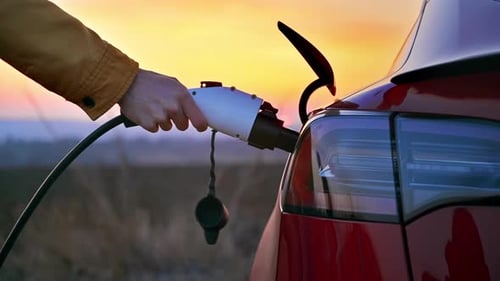 Nahaufnahme eines Mannes, der bei Sonnenuntergang ein Ladekabel in ein geparktes rotes Elektroauto steckt