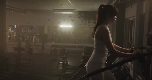 Young Woman Drinking Water and Exercising on Stair Climber in Dimly Lit Gym