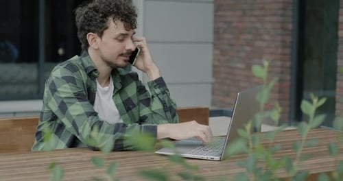 Middle Eastern Freelancer Speaking on Mobile Phone and Using Laptop Sitting in Outdoor Cafe in City