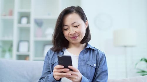 Close up Happy asian woman using smartphone while sitting on sofa in living room at home. Smiling