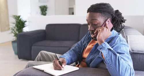 Homme afro-américain tenant un téléphone avec connexion d'appel, écrivant dans un cahier avec un stylo sur un canapé gris