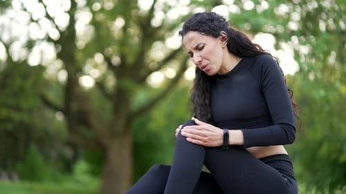 Close up. Young woman suffers from painful severe knee joint ache while sitting in urban city park.
