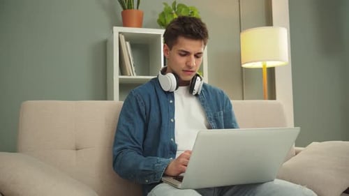 Junger Mann konzentrierte sich darauf, ein Problem zu lösen, während er in einem gemütlichen Innenraum an einem Laptop mit Kopfhörern arbeitete