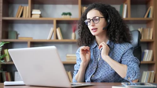 Confident woman in headset talking on video call using laptop sitting in home office.