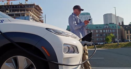 Man with Smartphone Waits While His Electric Car Charging at a Charging Station
