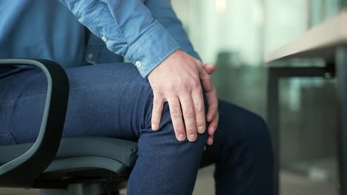 Close up. Male hands holding massaging knee while sitting at workplace in business office.