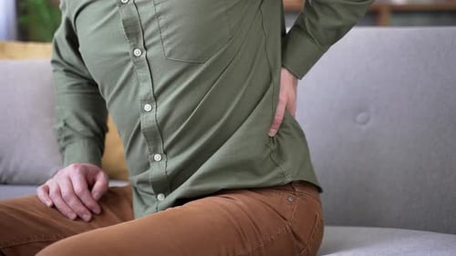 Close-up of male hands holding and massaging lower back while sitting on sofa at home. Man in shirt