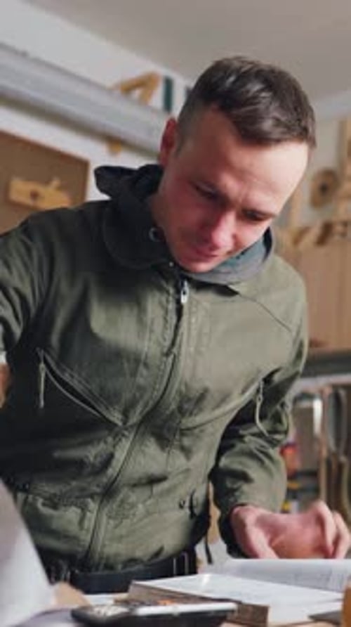 The Carpenter Works with Documents for a New Furniture Order in His Own Workshop A Young Male