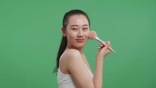 Asian Woman Wearing A White Camisole Rouging Her Cheeks And Smiling To Camera On Green Screen