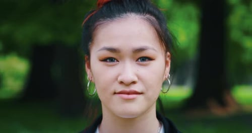 Slow motion of young asian woman is looking and smiling in camera on green city park background in