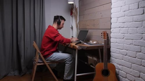 Young Man in Headphones Composing and Producing Music on a Laptop in a Cozy Home Studio with an