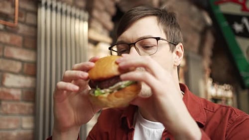Young Man Biting Burger Wearing Glasses Experiencing Culinary Delight in Rustic Restaurant with