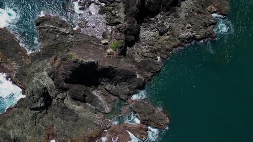 A stunning top-down drone shot captures the small rocky mountains along the shore, with the waves ge