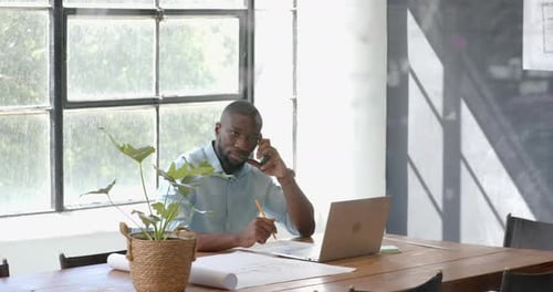 Talking on smartphone, man working on laptop and taking notes in office