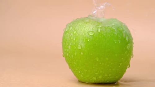Slow motion shot of water splashing on fresh green apple on beige background.