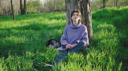 Woman Sits Against a Tree in a Green Field While Enjoying Sunlight and Has a Laptop Placed Beside