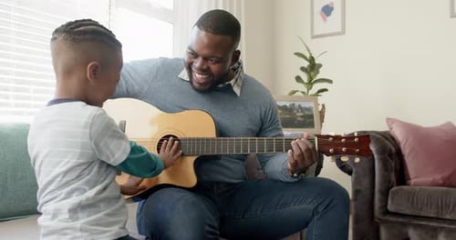 Happy african american father and son sitting on sofa and playing guitar, in slow motion
