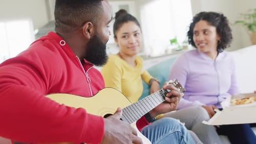 Happy, diverse female and male friends playing guitar and listening at home in slow motion