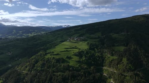 Aerial shot of lush green hills with a small village, surrounded by mountains and a partly cloudy sk