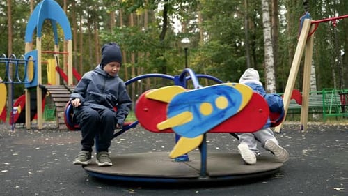 Two brothers in warm clothes are spinning on a carousel on a playground