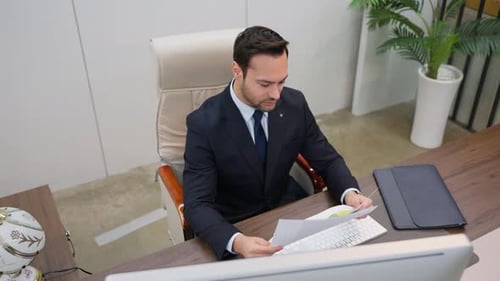 A businessman sits at a desk in an office reviewing paperwork documents or reports