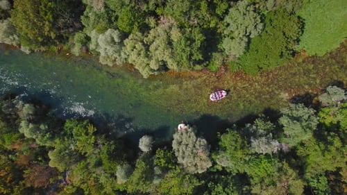 Rafts with people paddling down a clear, shallow river surrounded by dense, green forest. Aerial vie