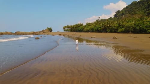 Woman in white dress walks on dark sandy beach with ocean waves in tropical Costa Rica