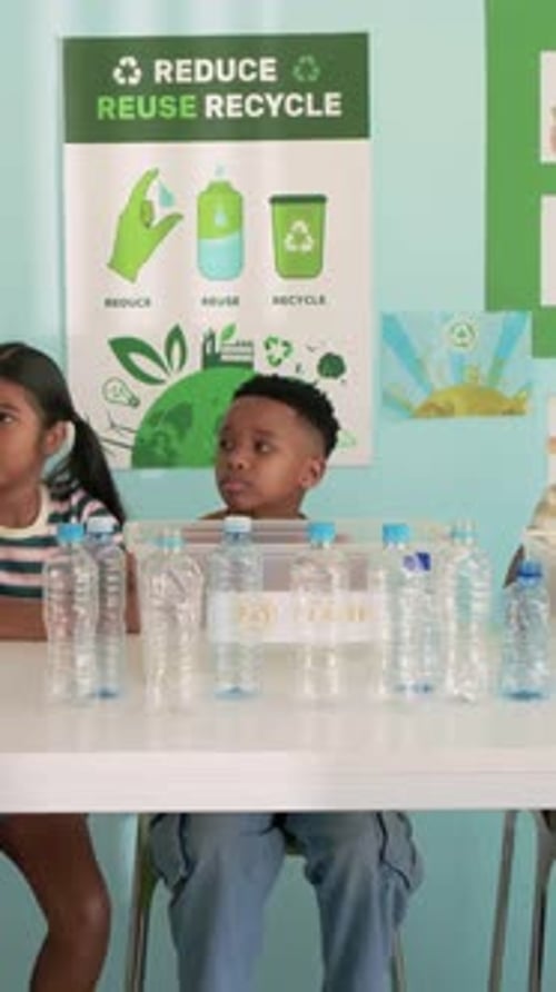 Vertical video: Adult cueing boy in blue pants leaning toward bottles in classroom for recycling