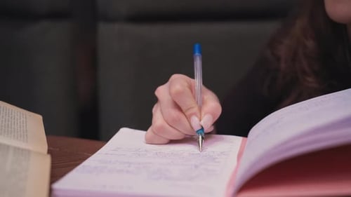 Close Up of Female Hand Writing in Pink Book Beside Open Brown Book