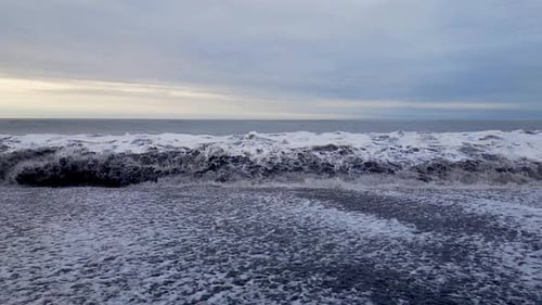 Surf waves forming white foam crashing onto a black sandy beach.