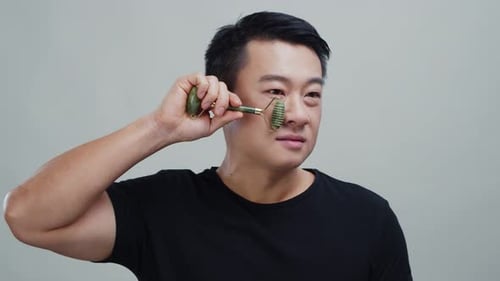 Asian Attractive Man Doing Morning Facial Routine Massaging Face with Green Jade Roller Isolated in