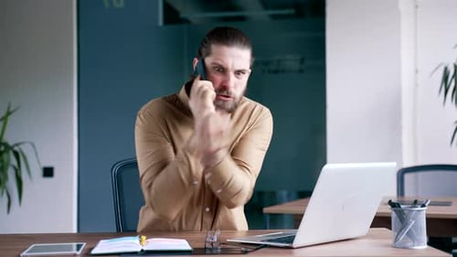 Angry businessman arguing talking on mobile phone sitting at workplace in office.